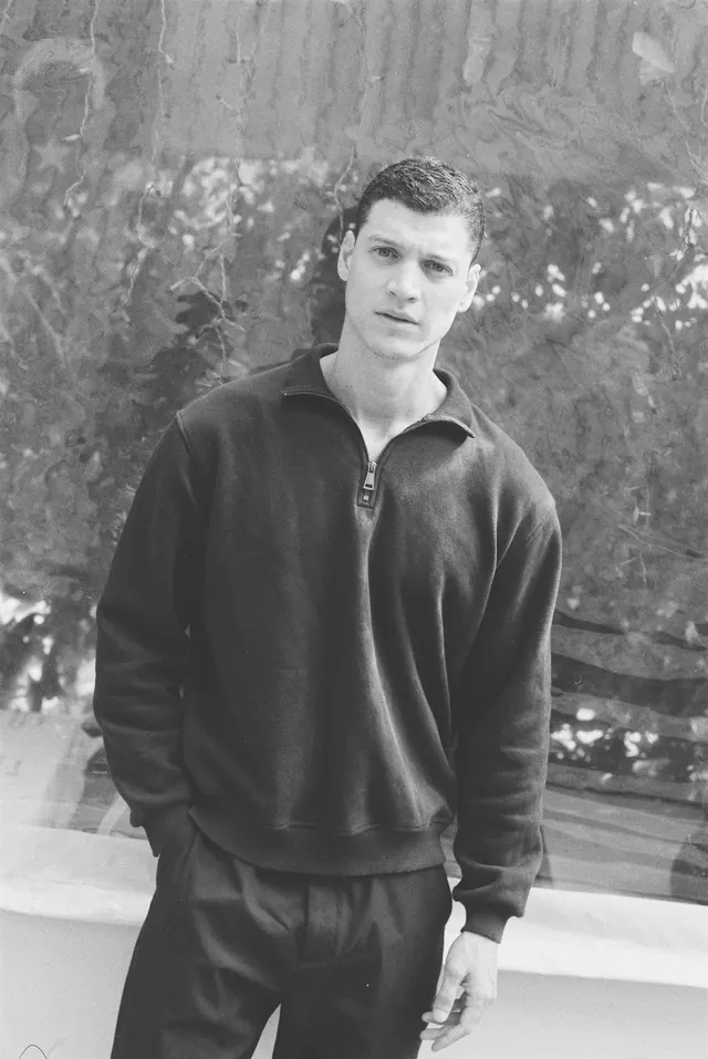 Black-and-white portrait in a dark sweater against a textured garden wall.