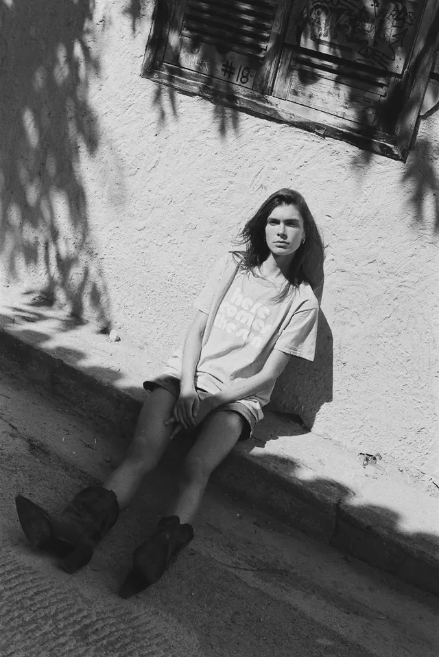 Black-and-white seated portrait beside a wall patterned with tree shadows.