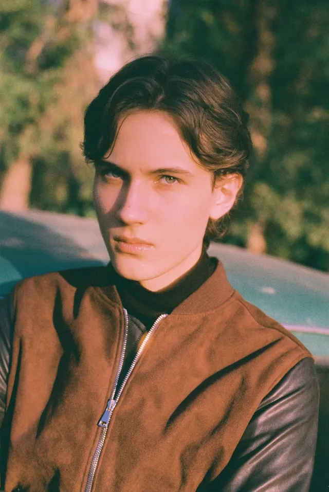 Close portrait in a brown jacket with warm foliage behind.