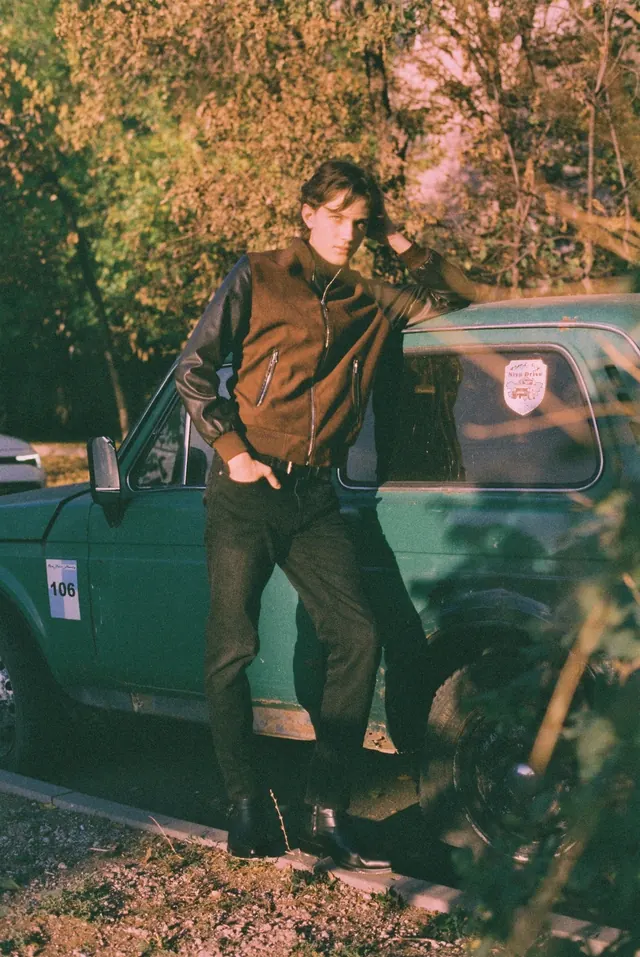 Full-length portrait in a brown jacket beside a blue car and autumn foliage.