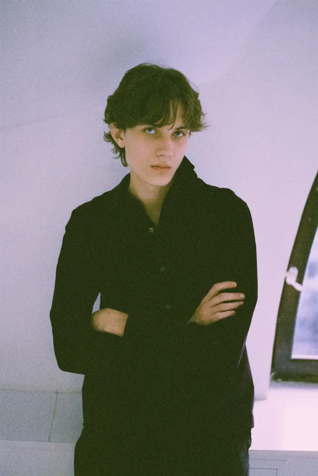 Portrait in black with folded arms against a pale lilac wall.