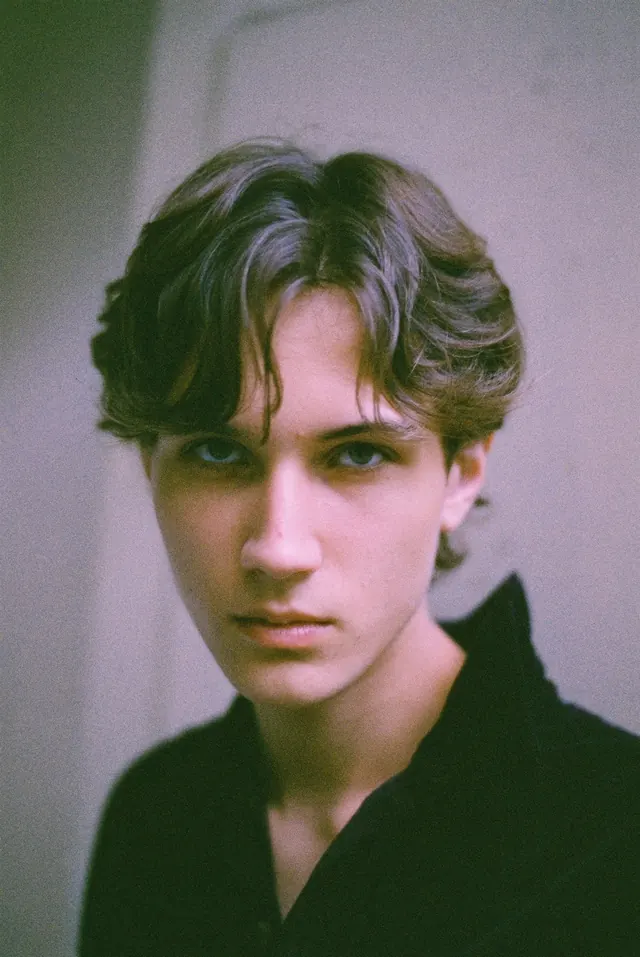 Close portrait in black, with a lilac wall softly blurred behind.