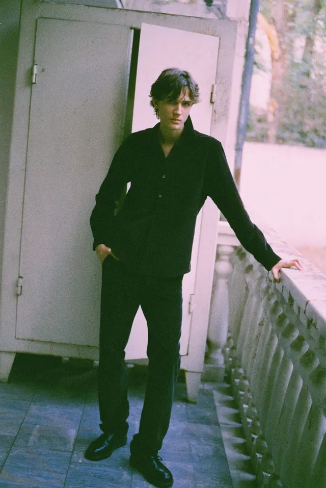 Full-length portrait in black beside an open door and balcony railing.