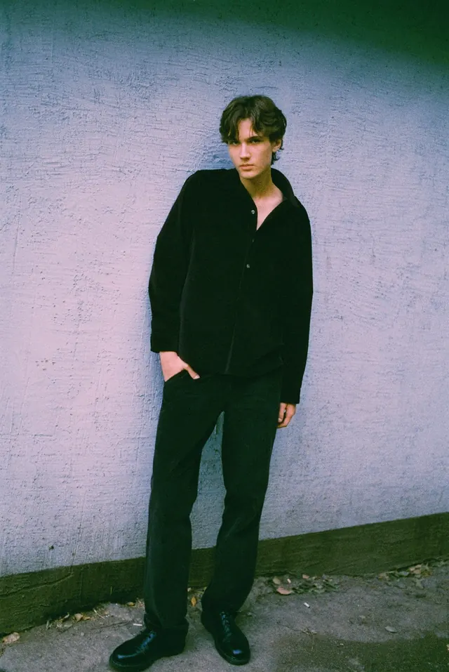 Full-length portrait in black against a lilac wall.