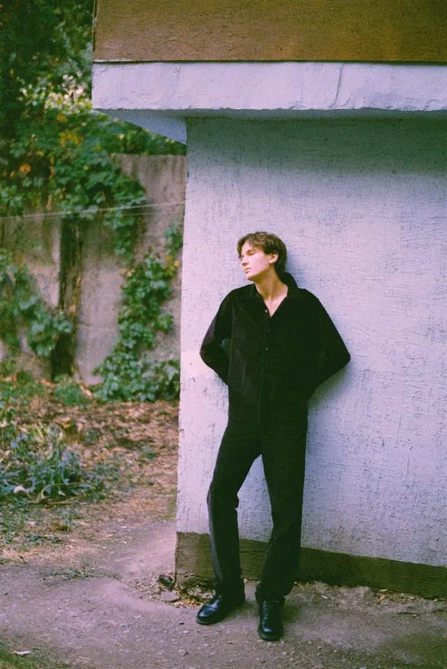 Full-length portrait in black at the corner of a pale building.