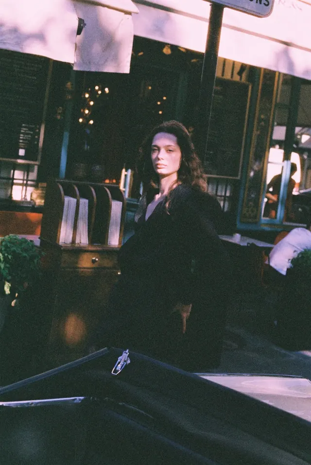 Portrait in black beside a cafe terrace and folding chairs.