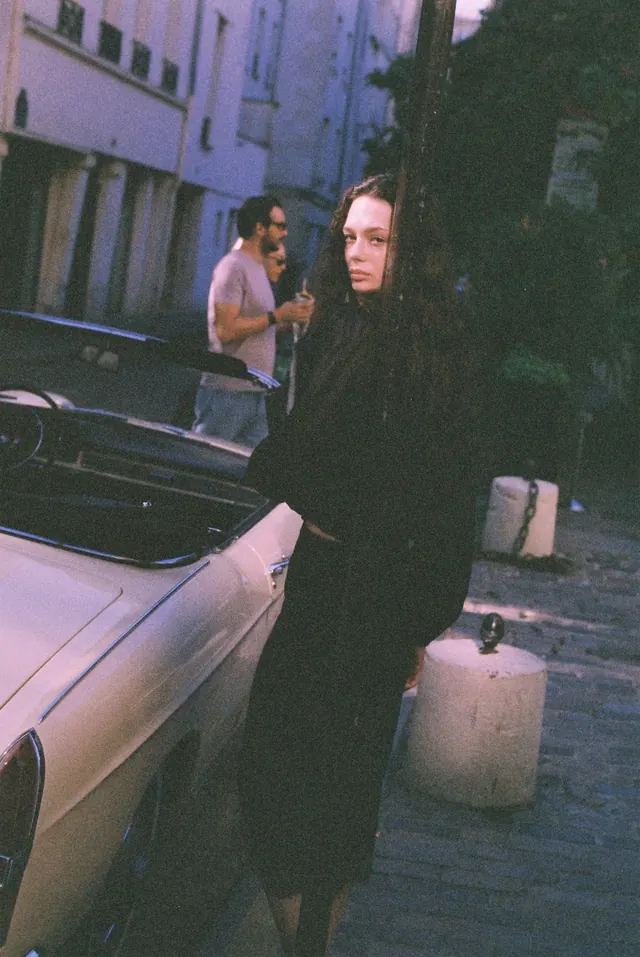Portrait in black beside a pale car on a shaded street.