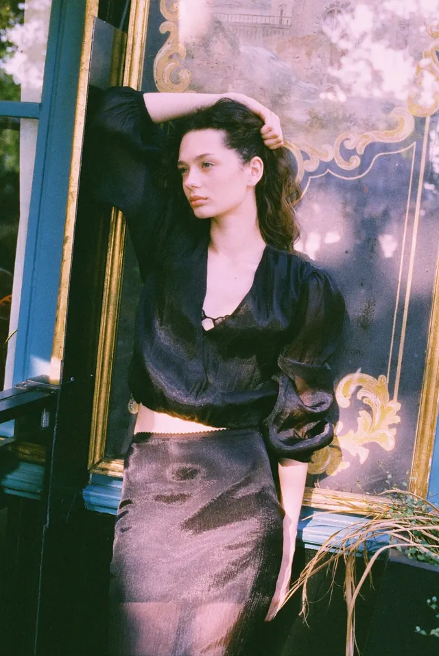 Portrait in a black blouse with one arm overhead beside an ornate cafe window.