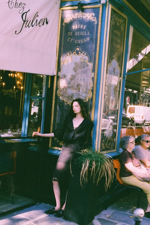 Full-length portrait beside an ornate cafe doorway and outdoor tables.