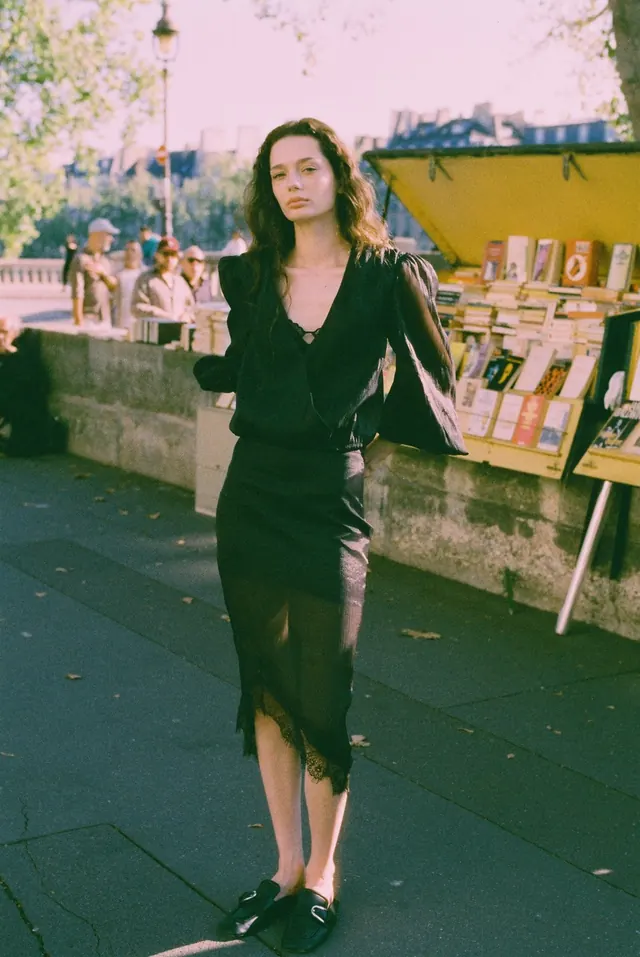 Full-length portrait in black beside riverside book stalls.