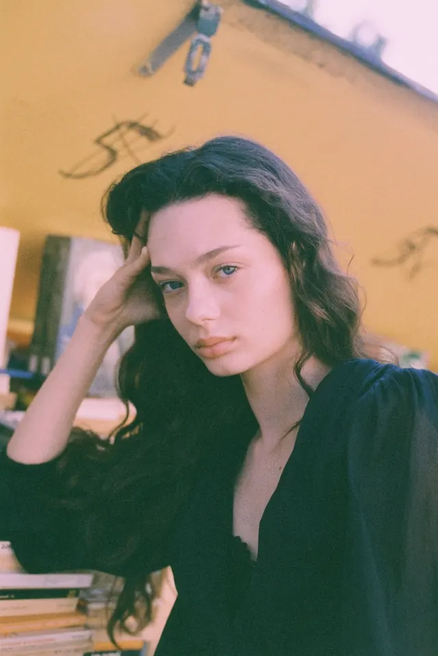 Portrait in a black blouse beside a yellow book stall, with one hand at the temple.