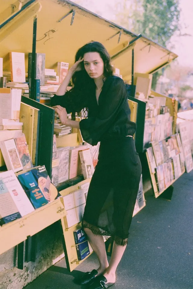 Full-length portrait in black among books at a yellow riverside stall.