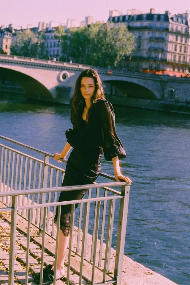 Portrait beside a river railing, with a stone bridge and city buildings behind.