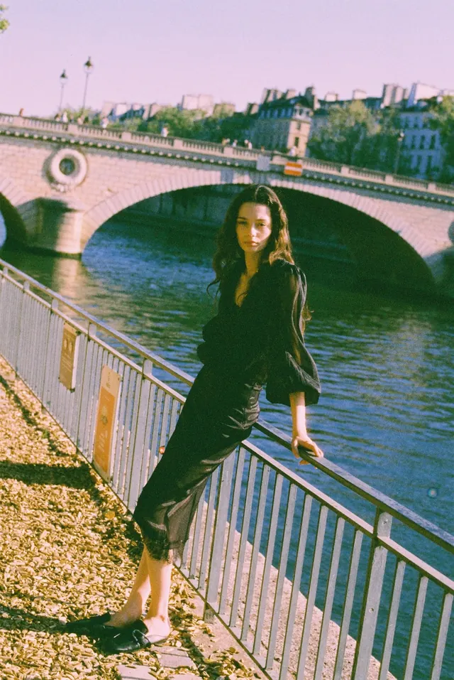 Full-length portrait leaning against a river railing beneath a stone bridge.