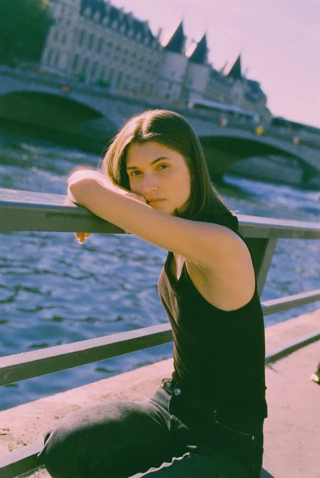 Sunlit portrait leaning on a river railing, with a stone bridge behind.
