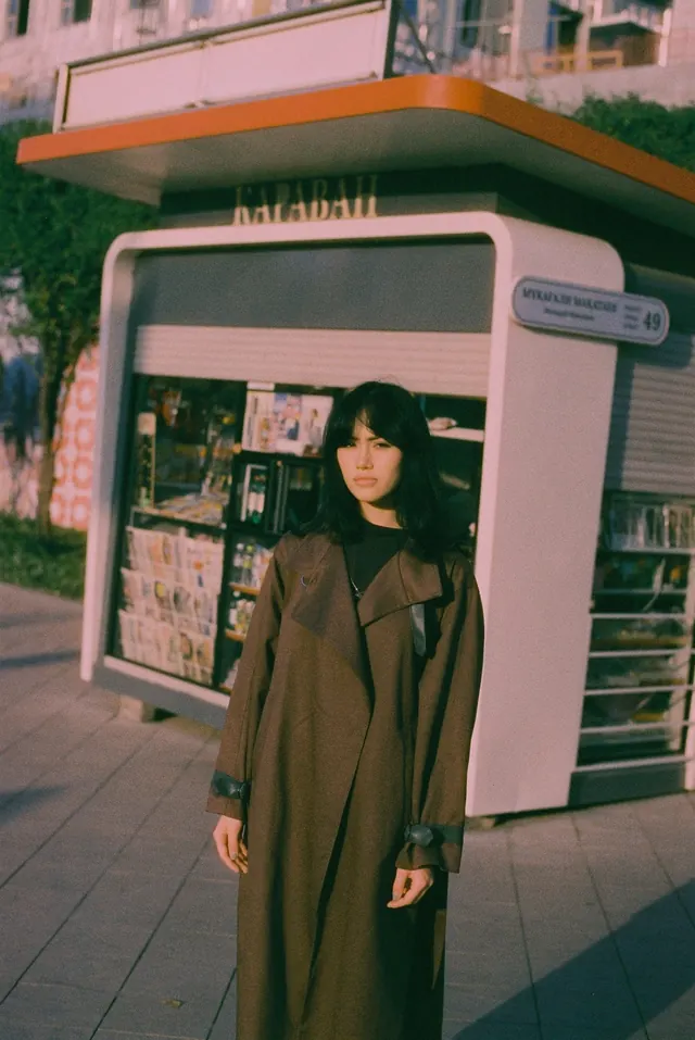 Portrait in a long brown coat before a street kiosk.