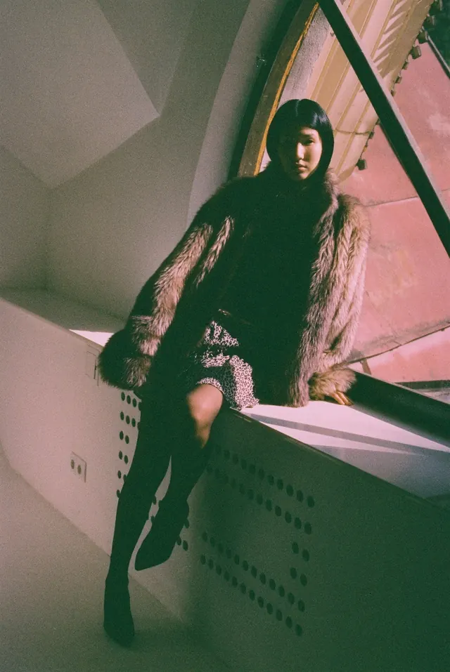Full-length portrait in a textured coat beside a sunlit window.