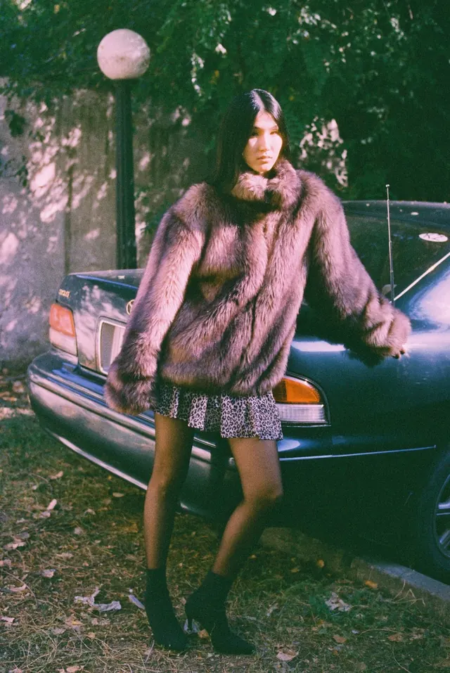 Full-length portrait in a textured coat beside a blue car and trees.